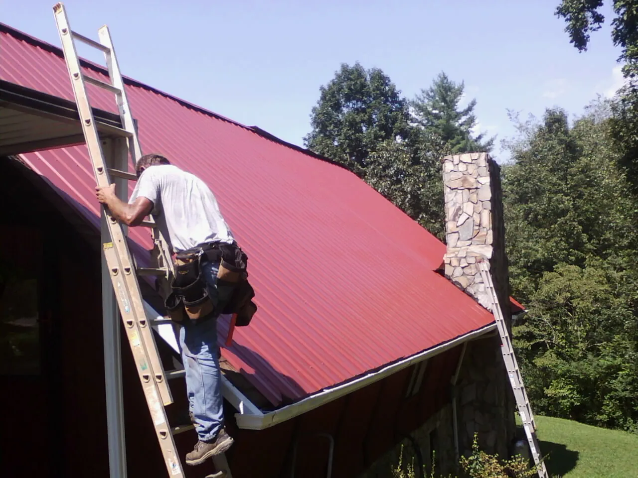 Roofer on a ladder beside a red metal roof and stone chimney — Soffit & Fascia Repair by Roofing Brothers in Mc Kean